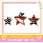 three wooden stars, each with a different design and color, arranged in a row. The stars are decorated with American flags and the word "America" written on them. The image also includes text that says "DIY WOOD PAINT KIT" and a heart symbol, suggesting that this is a kit for creating wooden stars.