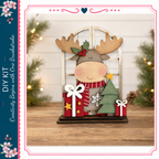 A wooden deer figurine with a Christmas tree and presents sits on a wooden surface, surrounded by holiday decorations and greenery.