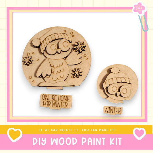 a DIY wood paint kit for creating a snow globe, with two wooden snow globe decorations in the shape of an owl. The kit includes instructions for creating the snow globe and the words "Owl Be Home for Winter" written on the kit.