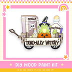 The image is a DIY wood paint kit featuring a wagon filled with Halloween-themed items, including a pumpkin, a witch's hat, and a spell book.