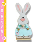 A wooden Easter bunny decoration kit, featuring a light brown bunny with pink ears and a light blue dress with buttons and a heart.