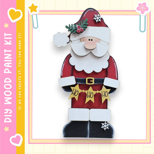 A wooden Santa Claus figure with a red suit, white beard, and hat, holding a "Ho Ho Ho" banner. The Santa Claus figure is set against a pink and yellow background with decorative elements such as hearts and snowflakes.
