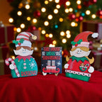 two wooden elf figurines and a sign with text, all placed on a red cloth against a blurred background with Christmas lights.
