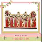 a large wooden sign with the word "BERRIES" written on it, surrounded by decorative elements such as flowers and stars. The sign is placed on a shelf, ready to be displayed.