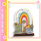 A colorful and whimsical wooden craft kit featuring a rainbow, a pot of gold coins, and a hat, all set against a pink and yellow background.