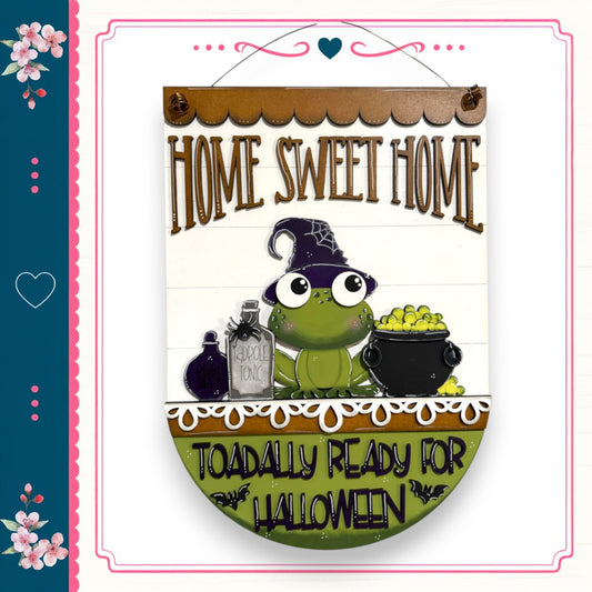 a sign with a frog wearing a witch's hat and holding a cauldron, accompanied by the text "Home Sweet Home" and "Toadally Ready for Halloween".