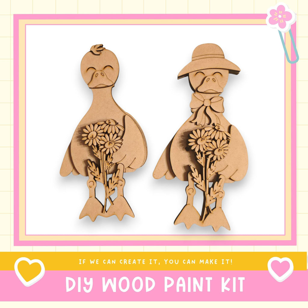 two wooden bird figurines, one with a flower in its beak and the other holding a flower in its hand. The image is framed with a pink border and includes text that reads "DIY WOOD PAINT KIT" and "If we can create it, you can make it!"