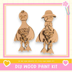 two wooden bird figurines, one with a flower in its beak and the other holding a flower in its hand. The image is framed with a pink border and includes text that reads "DIY WOOD PAINT KIT" and "If we can create it, you can make it!"