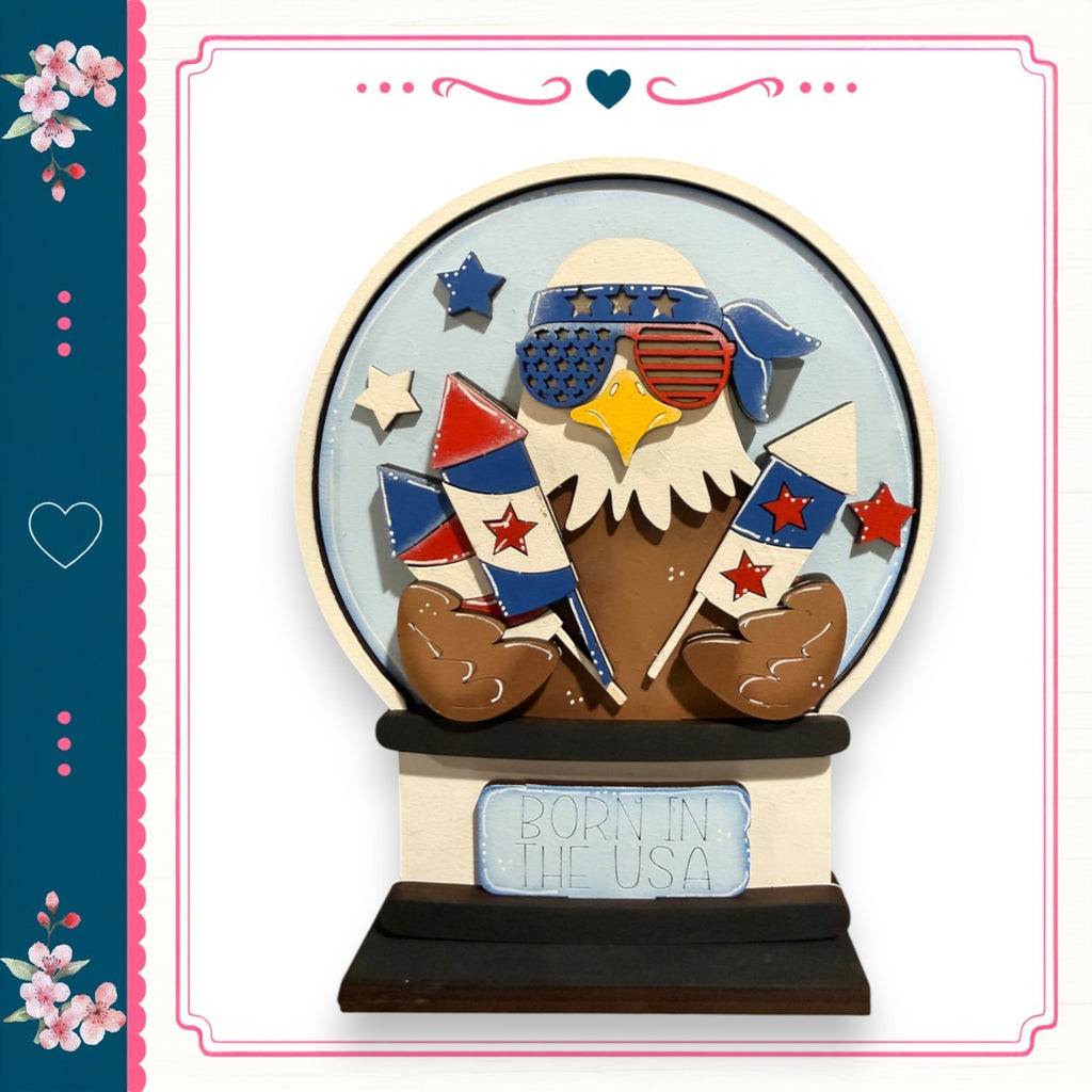 A decorative snow globe with an eagle wearing sunglasses and holding fireworks, surrounded by stars and flowers, with the text "Born in the USA" on the base.