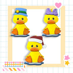A collage of three cheerful yellow duck figures wearing hats and bow ties, set against a grid background with colorful stickers.