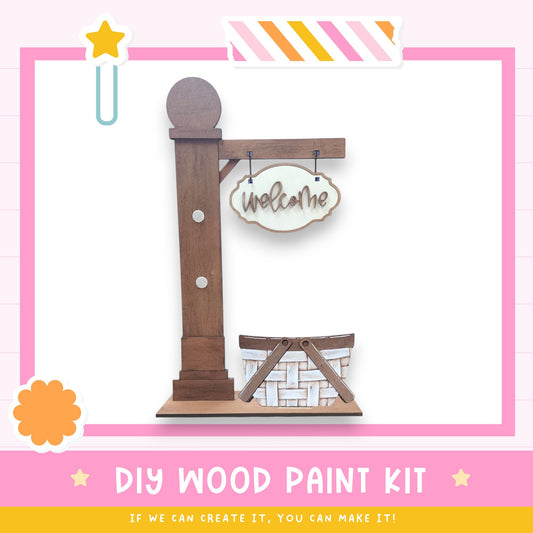 a DIY wood paint kit, including a wooden sign with the word "Welcome" and a basket, all set against a pink and white background.