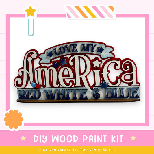 The image is a colorful and playful DIY wood paint kit for creating a patriotic sign or decoration. It features the words "I Love My America" in a decorative font, with red, white, and blue colors. The kit appears to be designed for children, as indicated by the playful design and the use of bright colors.