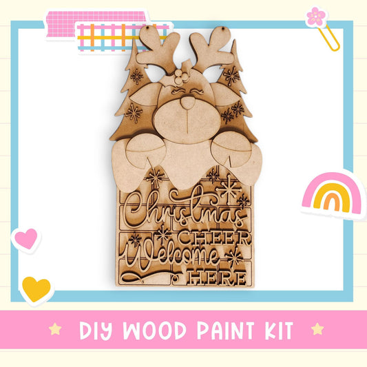 a DIY wood paint kit for creating a Christmas-themed sign. The kit includes a wooden sign with a reindeer and snowflake design, as well as a wooden sign with a "Christmas Cheer Welcome Here" message. The kit is presented in a pink and blue frame, and the text "DIY WOOD PAINT KIT" is displayed at the bottom of the image.