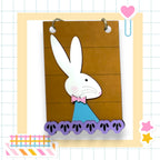 A paper craft of a white rabbit wearing a blue bow tie, with a brown background and purple paw prints, is displayed in a frame.
