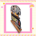 A wooden bat decoration with a bat and a rainbow-colored background, surrounded by a pink and yellow checkered border.