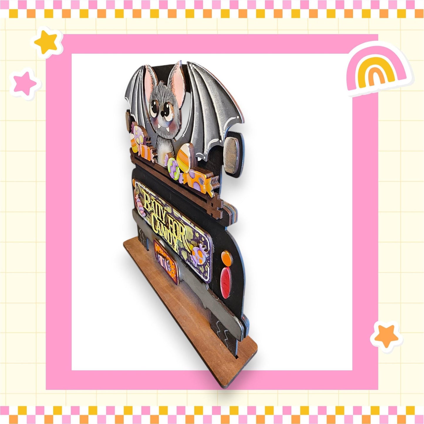 A wooden bat decoration with a bat and a rainbow-colored background, surrounded by a pink and yellow checkered border.