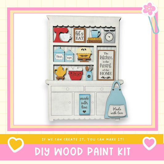 The image is a DIY wood paint kit featuring a wooden cabinet with various kitchen-themed items such as a mixer, measuring cups, a clock, and a sign that says "The kitchen is the heart of the home".