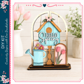 A wooden house-shaped decoration with a "Hello Spring" sign, flowers, and a watering can, placed on a wooden surface.