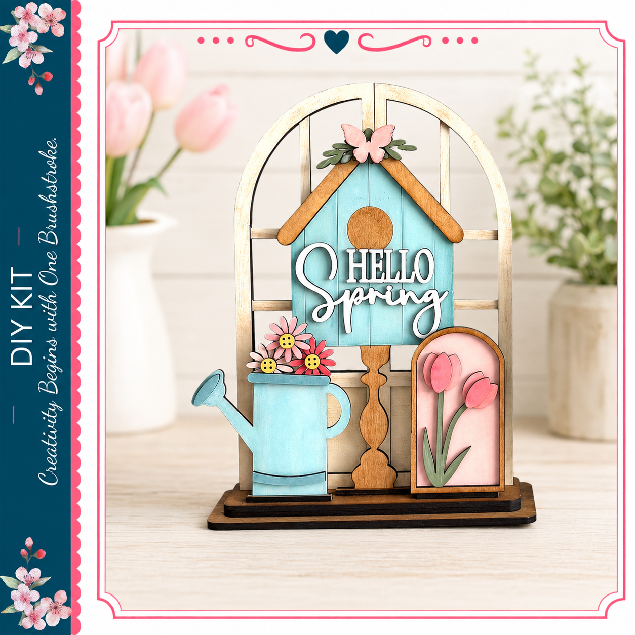 A wooden house-shaped decoration with a "Hello Spring" sign, flowers, and a watering can, placed on a wooden surface.