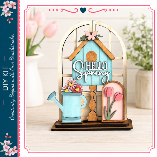 A wooden house-shaped decoration with a "Hello Spring" sign, flowers, and a watering can, placed on a wooden surface.