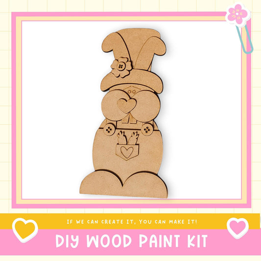 a DIY wood paint kit, with a wooden gnome figure as the main focus. The gnome is wearing a hat with a flower and has a heart-shaped bow around its neck. The kit appears to be made of wood and is designed for creating wooden figures.