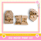 three wooden ornaments with various designs, including a Santa Claus figure, a pair of boots, and a tag with the word "Merry". The image also includes text that says "DIY WOOD PAINT KIT" and "IF WE CAN CREATE IT, YOU CAN MAKE IT!"