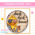 a wooden sign with a duck wearing sunglasses and holding fireworks, accompanied by the text "duck, duck boom!"
