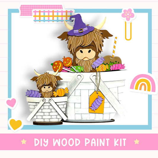 The image is a colorful illustration featuring two cartoon cow characters, one wearing a witch's hat, sitting in a basket filled with various Halloween-themed items such as candy, pumpkins, and a rainbow. The image also includes a DIY wood paint kit and the text "DIY WOOD PAINT KIT" in the bottom right corner.