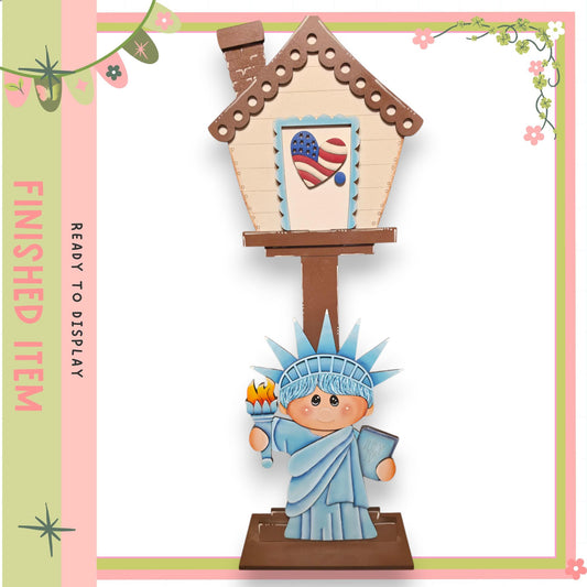 A wooden house with a painted American flag on the front, featuring a small statue of the Statue of Liberty in the foreground.