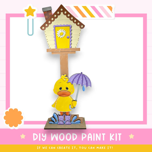a wooden house with a yellow door and a yellow flower on the door, along with a yellow duck holding a purple umbrella. The image also includes a pink and white striped border, a star, and the text "DIY WOOD PAINT KIT" and "IF WE CAN CREATE IT, YOU CAN MAKE IT!"
