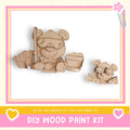 two wooden toy bears, one larger and one smaller, sitting together and smiling. The image is framed with a pink border and includes text that reads "DIY WOOD PAINT KIT" and "If we can create it, you can make it!"