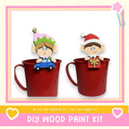 two elf figurines, one sitting in a red mug and the other in a red mug, both wearing Santa hats. The image also includes text that says "If we can create it, you can make it!" and "DIY WOOD PAINT KIT".
