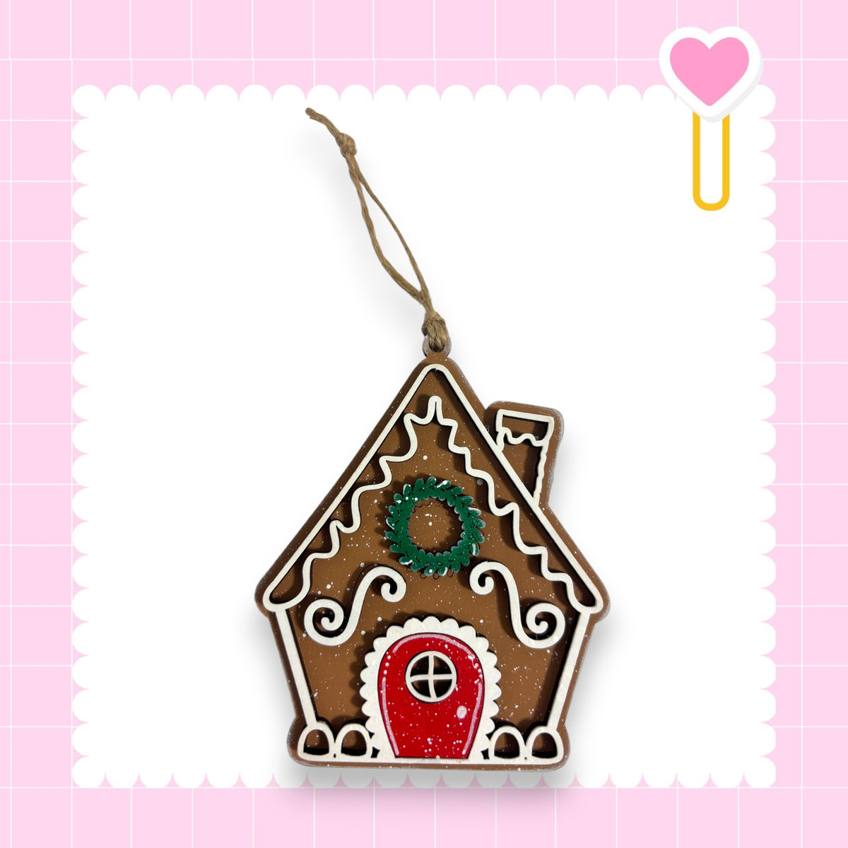 A gingerbread house ornament with a wreath on top, hanging from a string against a pink background with a scalloped edge.