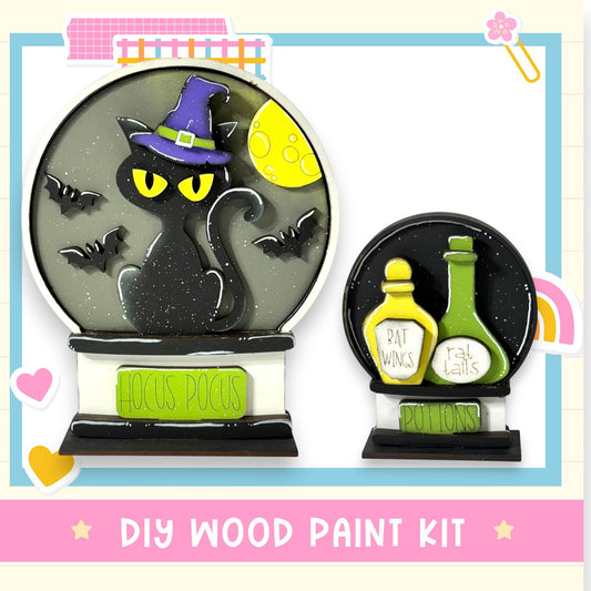 a Halloween-themed DIY wood paint kit, including a black cat wearing a witch's hat, a glass snow globe, and two bottles labeled "BAT WINGS" and "POTIONS".