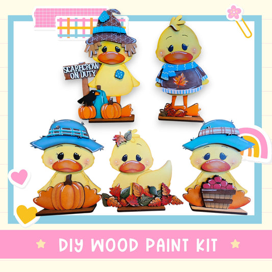 The image is a collage of five wooden duck figurines, each wearing a different hat and holding various fall-themed items such as pumpkins and apples. The text "DIY WOOD PAINT KIT" is displayed at the bottom of the image.