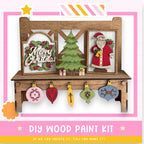 The image is a DIY wood paint kit featuring a wooden shelf with a Christmas tree, a framed image of Santa Claus, and various ornaments hanging from the shelf.