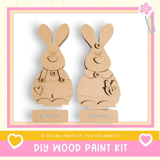 two wooden rabbit figures, each with a heart and a bow, standing on a wooden base. The text below the image reads "DIY WOOD PAINT KIT" and "If we can create it, you can make it!"