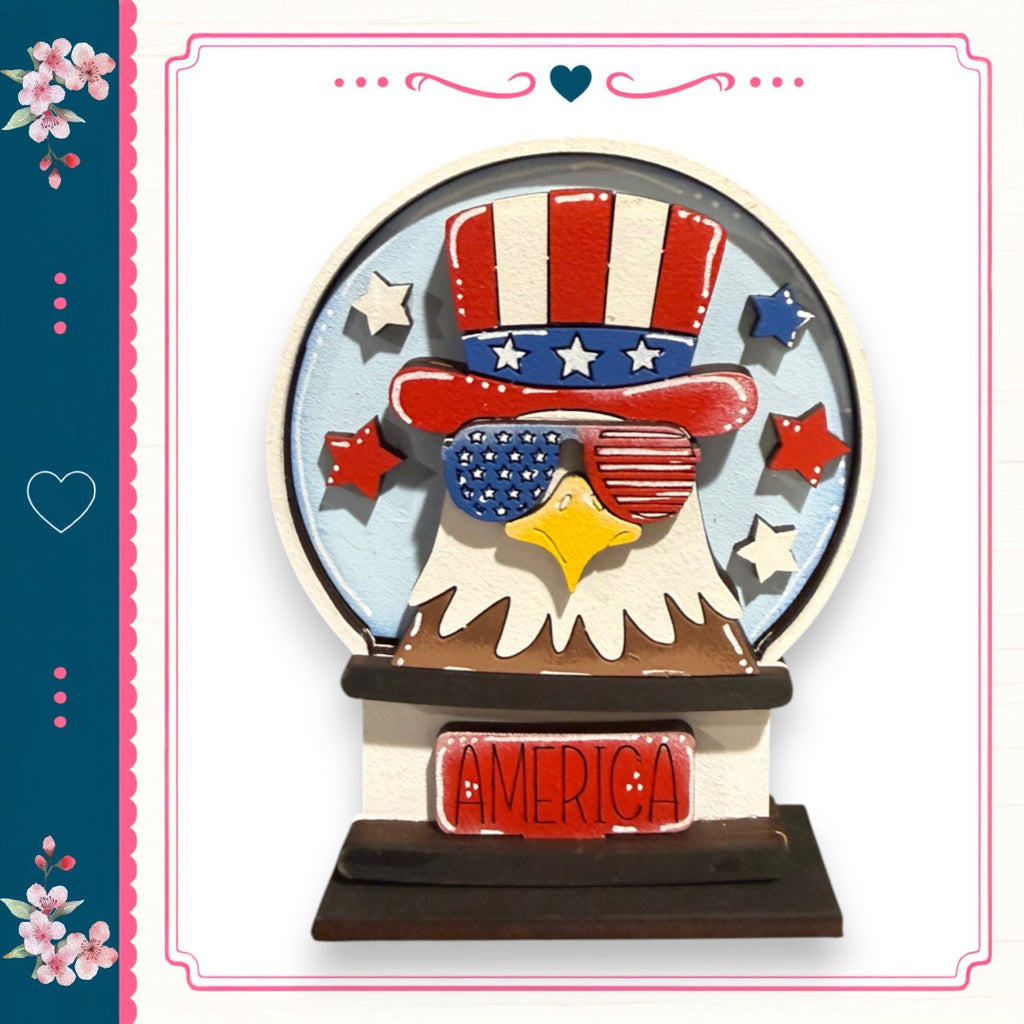 A wooden snow globe with an eagle wearing sunglasses and an American flag hat, surrounded by stars and flowers.