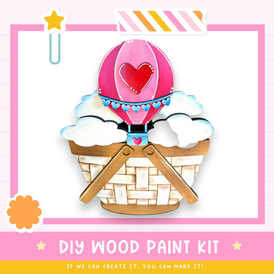 The image is a colorful and whimsical DIY wood paint kit featuring a hot air balloon, a basket, and clouds. The kit appears to be made of wood and is designed for painting.