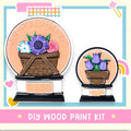 The image is a DIY wood paint kit featuring two wooden baskets with flowers, one larger and one smaller, set against a light blue background with a pink border.