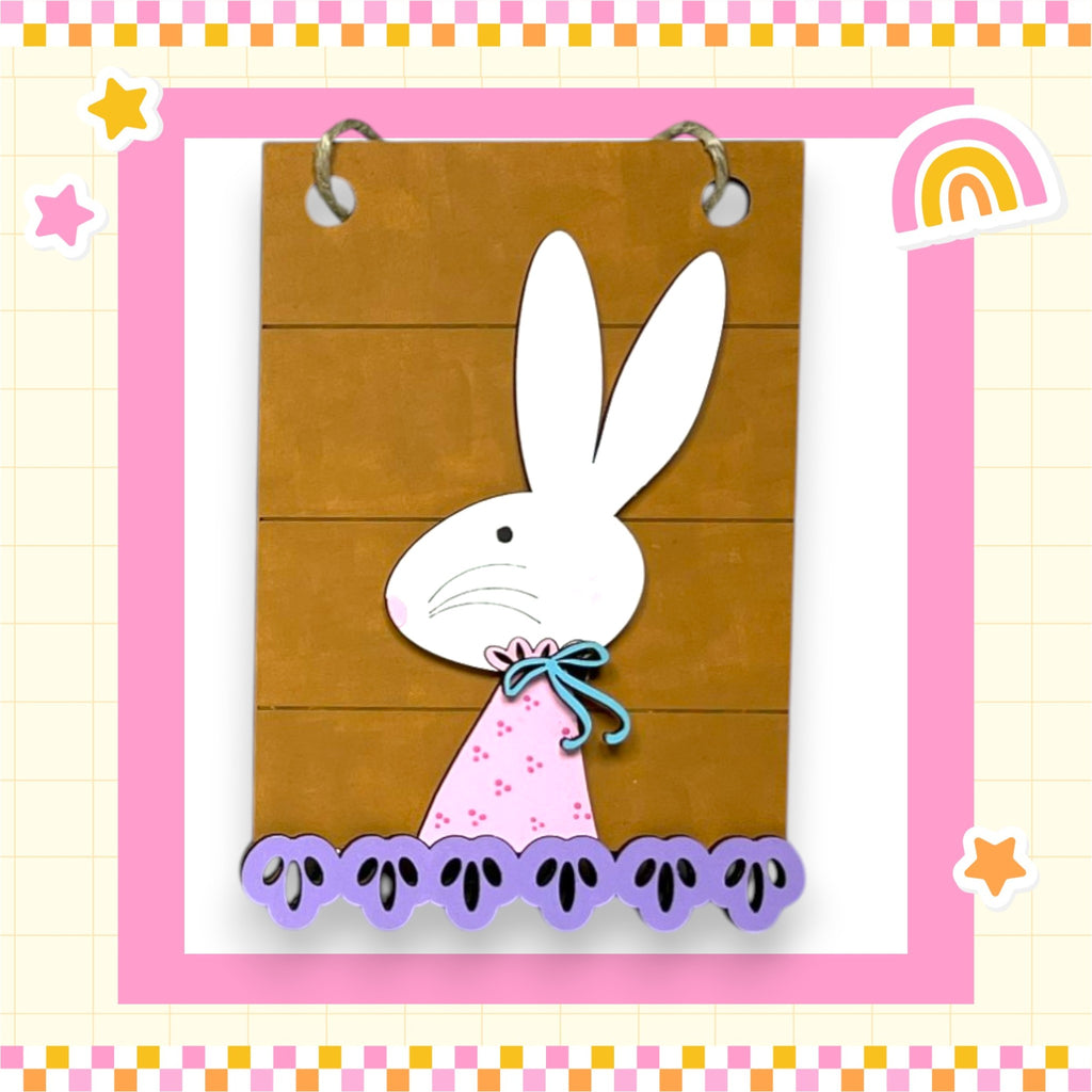A wooden sign with a white rabbit figure on it, surrounded by purple flowers and a pink bow.
