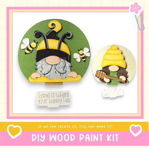 two wooden gnome figurines, one larger and one smaller, with bee and honeycomb decorations. The larger gnome is wearing a yellow hat and holding a honeycomb, while the smaller gnome is holding a wooden spoon. The image also includes a note that says "Home is where your honey bee" and a DIY wood paint kit, suggesting that the gnomes can be created using the provided kit.