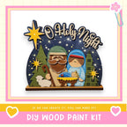 A DIY wood paint kit for creating a nativity scene with a wooden frame, featuring a figure of the baby Jesus, Santa Claus, and Mary, surrounded by stars and greenery.