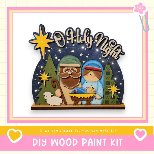 A DIY wood paint kit for creating a nativity scene with a wooden frame, featuring a figure of the baby Jesus, Santa Claus, and Mary, surrounded by stars and greenery.