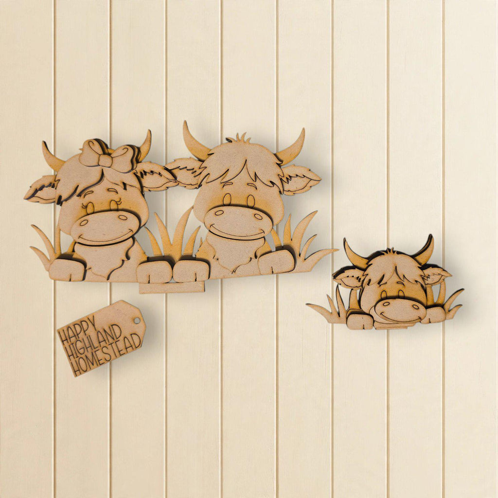 A wooden cutout of three cows, two of which are larger and the third one is smaller, sitting in the grass. There is also a small wooden tag with text attached to the wall.