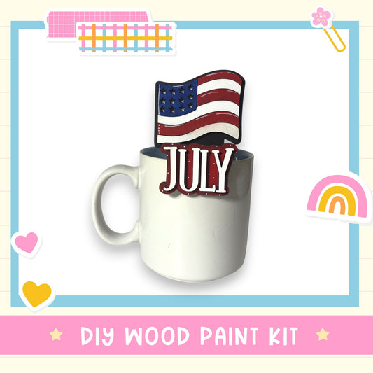 A white mug with a patriotic American flag design on top, along with the word "JULY" written on it. The mug is placed on a white background, and there is a DIY wood paint kit visible in the image.