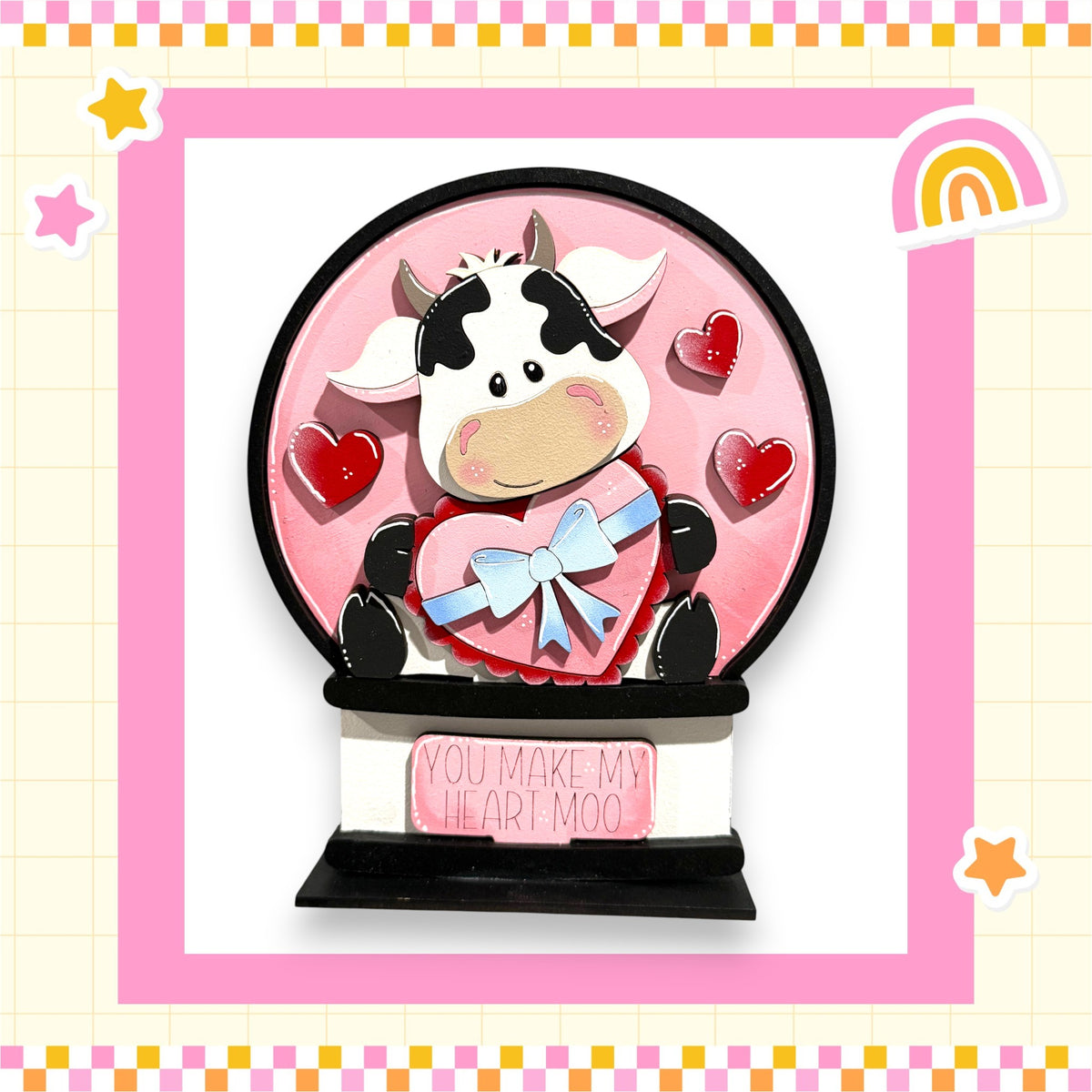 A heart-shaped ornament with a cow figure inside, surrounded by hearts and a pink background. The ornament has a sign that says "You make my heart moo".