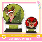 two wooden snow globes, each containing a reindeer. The snow globes are set against a green background, and the text below the image encourages viewers to create their own DIY wood paint kit.