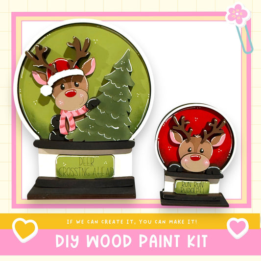 two wooden snow globes, each containing a reindeer. The snow globes are set against a green background, and the text below the image encourages viewers to create their own DIY wood paint kit.