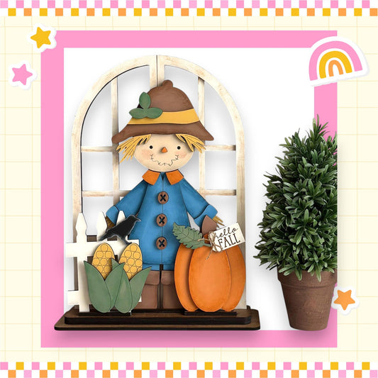 A wooden scarecrow figure holding a sign that says "Hello Fall" stands in front of a window, with a potted plant and cornucopia nearby.
